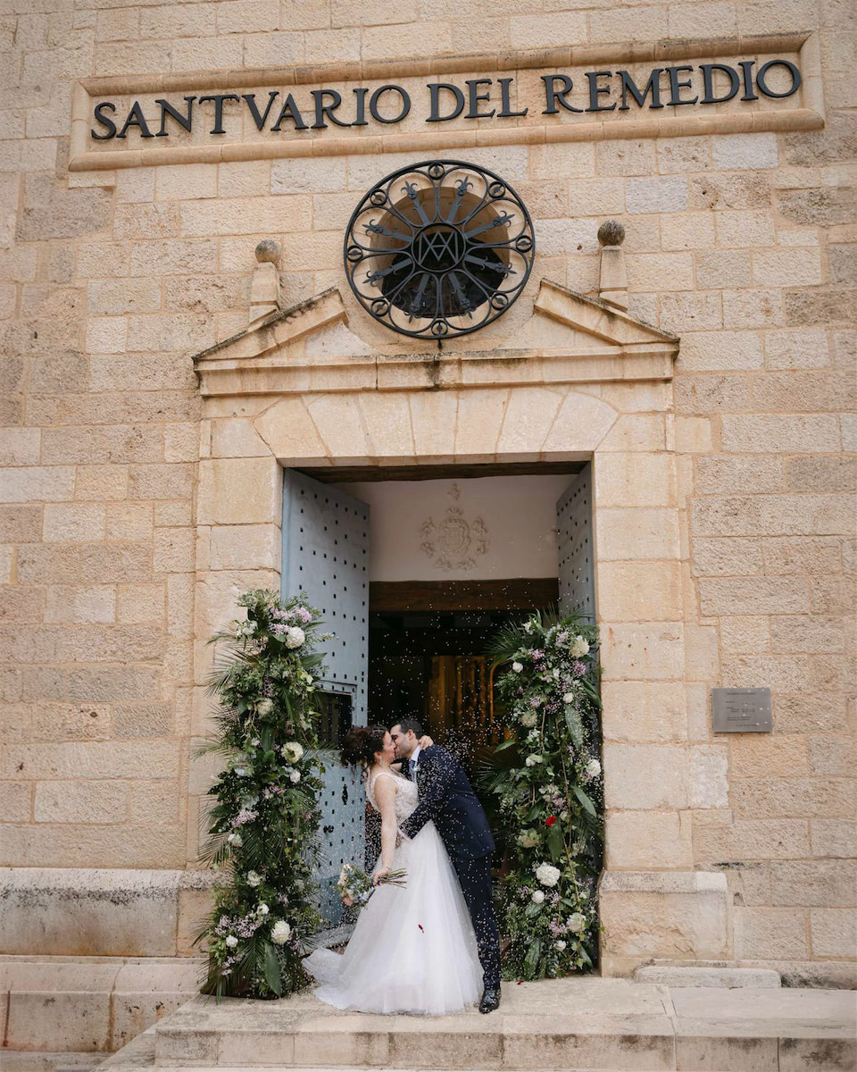 Los novios se besan a la salida de la Ermita del Remedio de Utiel mientras sus invitados les lanzan arroz, con la fachada de la ermita al fondo.