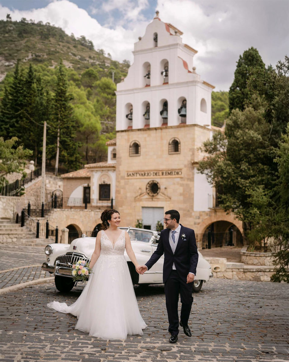 Novios recién casados caminando de la mano junto a la Ermita del Remedio de Utiel, con un coche clásico al fondo.